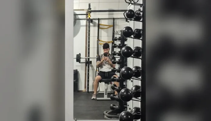 Michel Silva sentado num banco de musculação na academia, em frente à arara de kettlebells
