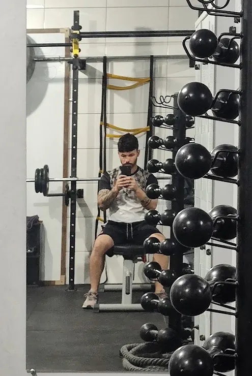 Michel Silva sentado num banco de musculação, em frente à arara de kettlebells da academia